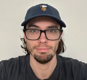 Man with small beard wearing clear eyeglasses and a small hat.